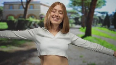 Sarışın, neşeli bir şekilde gülümseyen, güneşli bir park ortamında kollarını açmış, açık havada mutluluğu ve özgürlüğü gösteren genç bir kadın..