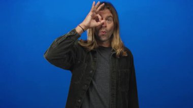 Sarışın, uzun saçlı bir adam mavi stüdyo duvarına ve büzüşen dudaklara karşı tamam işareti tutuyor; merak keşfi oyunculuğu.