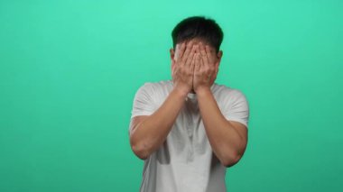 İzole edilmiş yeşil arka plan duvarındaki genç Çinli adam, elleri ile gözleri örtüp açarken neşeli, neşeli bir anı videoda yakalıyor..