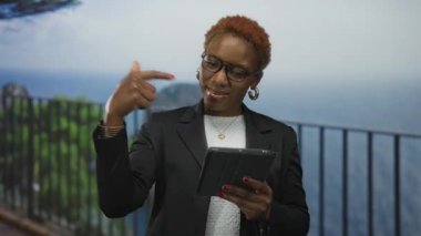 Genç Afro-Amerikalı kadın, deniz kenarındaki trabzan kenarında elinde tabletle parmak işareti yapıyor; davetiye bağlantısı.