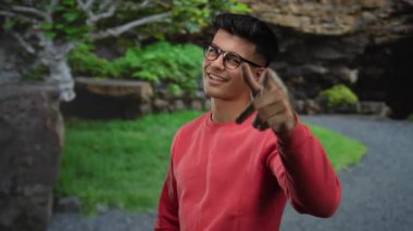 Gözlüklü ve kırmızı kazaklı genç adam, parlak bir günde yemyeşil bir parkta güvenle dikiliyor..