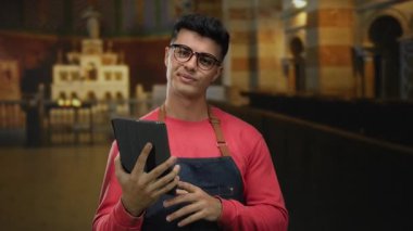 Gözlüklü genç adam kilisenin içinde tablet tutuyordu. Süslü mimari ve kemerlerle, günlük önlükler giyiyordu. Kapalı alanda yansıtıcı bir şekilde dikilirken kendine güvenini gösteriyordu..