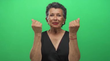 Gri saçlı yaşlı kadın, parmaklarıyla yeşil bir zemine işaret ediyor, iyimserlik ve başarı ifadesi gösteriyor, zenginlik ve pozitifliği vurguluyor..