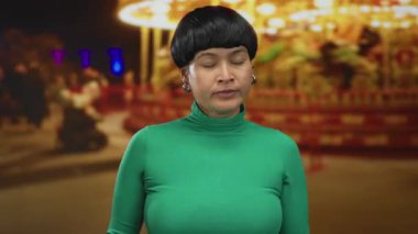 Canlı bir gece fuarında yeşil kazak giyen, arka planda parlak ışıklar ve bulanık insanlar olan bir kadın açık havada canlı bir atmosfer yaratıyor..