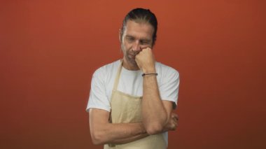 Sarışın, önlüklü bir adam elleriyle yanağına dokunuyor ve turuncu stüdyoda kollarını kavuşturuyor..