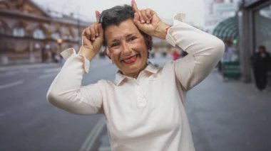 Gri saçlı yaşlı kadın sokak ortamında boynuz hareketi yapıyor, gülümsüyor ve açık havada neşeli bir sahne yaratıyor..
