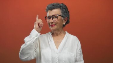 Gri saçlı, gülümseyen ve turuncu arka planda kafasını gösteren, gözlüklü, beyaz gömlekli, düşünceli bir ifade ve kendine güvenen bir tavır sergileyen son sınıf öğrencisi kadın..