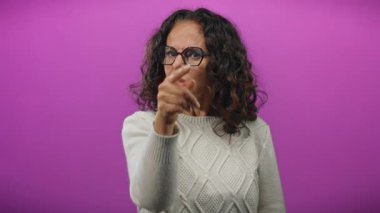 Canlı mor bir arka plana karşı ciddi bir ifadeyle kameraya doğru hareket eden bir kadın, sahnede aktarılan etkileşimi ve ruh halini vurguluyor..