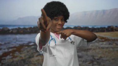 Steteskoplu kadın doktor, gülümserken güneşli kumsalda kamerayı işaret ediyor..