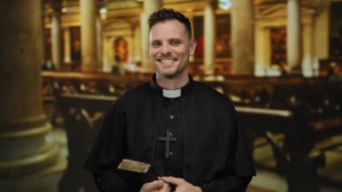 Kilise ortamında, elinde İncil, inanç ve dini bağlılık simgesi olan siyah cübbe ve çapraz kolye ile güvenle gülümseyen bir rahip..
