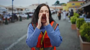 Turuncu yelekli bir kadın kalabalık bir sokakta gülümsüyor ve bağırıyor. Canlı bir şehir bölgesinde gönüllü olarak çalışıyor..