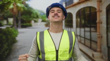 Yansıtıcı yelek ve kasketli genç İspanyol bir adam, şehir inşaat temalarını somutlaştıran bir caddede güven içinde duruyor..