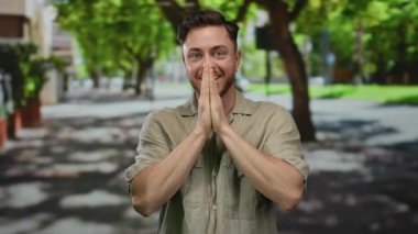 Sakallı genç bir adam yazın güneşli bir sokakta arka planda yeşil ağaçlarla gülümsüyor, rahat bir gömlek giyiyor ve neşeli bir ifade sergiliyor.