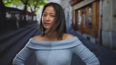 Şehir caddesinde ciddi bir ifadeyle duran genç bir kadın, mavi bir bluz giyiyor, arka planda bulanık ağaçlarla şehir manzarası sergiliyor..