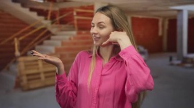 Kapalı inşaat alanında pembe gömlekli, el işaretli, tuğladan duvarları olan bir kadın ev ortamında neşeli ve kendinden emin bir ruh hali yaratıyor..