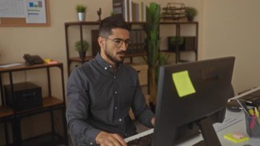Ofiste bilgisayarla çalışan, sırtı ağrıyan, bitkilerle ve ofis malzemeleriyle çevrili, gözlüklü ve siyah gömlekli, odaklanmış ve İspanyol asıllı bir adam..