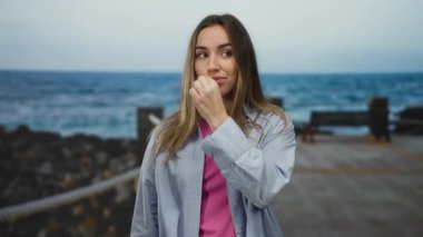 Sarı saçlı genç bir kadın, arka planda okyanus manzaralı bir deniz kenarında dikilirken ağzını kapatıyor. Üzerinde bir gömlek ve açık havada pembe bir üst var..