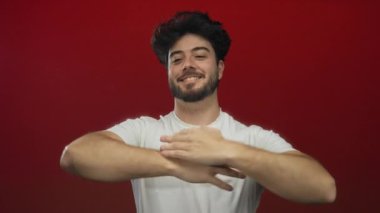 Kolları açık gülümseyen genç bir adam canlı kırmızı bir arka planda neşeli bir hoş geldin jesti sergiliyor sakallı ve günlük beyaz bir tişört giyiyor..