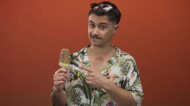 İzole edilmiş turuncu arka plan duvarında dondurmayı işaret eden İspanyol genç turist kafasında tropikal bir gömlek ve güneş gözlüğü ile merakla gülümsüyor..