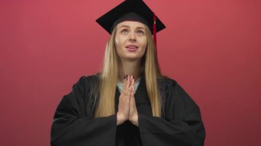 Mezuniyet elbisesi giymiş, kırmızı duvarın karşısında dua eden kadın, elleri bağlı, düşünceli bakışlarıyla akademik başarıyı ve gelecek umutlarını kutlayan umut dolu bir bakış atıyor..