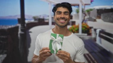 Adam göğsünün önünde beyaz kurdeleli yeşil hediye kutusunu taştan bir duvarın yanında güneşli bir deniz kenarında tutuyor..