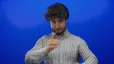 Göğsünün önünde kollarını mavi arka planda birleştiren sakallı, genç İspanyol bir adam. Onaylamadığını belirtiyor ve çizgili gömlekle jest yapıyor..