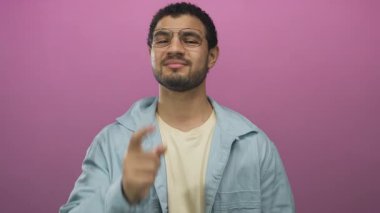 Stüdyoda pembe duvarlı, gözlüklü ve kot gömlekli bir adam kamerayı işaret ediyor..