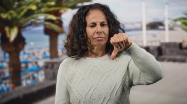 Kıvırcık saçlı kadın deniz kenarında baş parmağıyla duruyor sahilde hayal kırıklığı yaşıyor, açık havada süveter giyiyor..