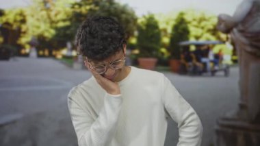 Güneşli şehir parkında gözlüklü ve çatık kaşlı İspanyol genç adam diş ağrısı için yanak yanağa bastırıyor; acı sıkıntı ıstırabı.