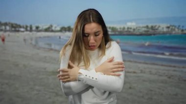 Deniz kenarında soğuk hisseden genç bir kadın manzaralı, ıslak, okyanus kıyısında beyaz bir kazak giyiyor. Etrafı bulanık insanlarla çevrili. Açık hava serin.