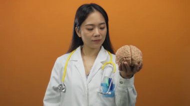 Turuncu arka planda steteskoplu beyin modelini tutan kadın doktor, tıp eğitimi ve sağlık uzmanlığını simgeliyor