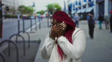Gözlüklü ve kırmızı örgülü bir kadın, şehir caddesinde elleriyle yüzünü örterek, gündüz vakti korku ve endişe ifade ediyor..
