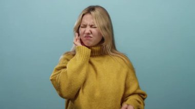 Dişi ağrıyan, sarı süveter giyen, izole edilmiş mavi arka planda acı çeken, yüzünde bir ifade ve yanağında bir el olan bir kadın..