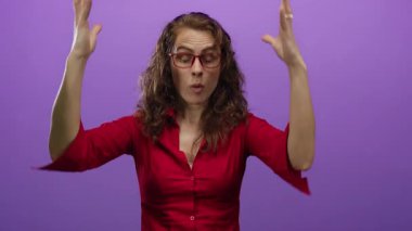 İzole edilmiş mor bir duvara karşı hareketli bir kadın, kırmızı gömlek ve gözlük giyiyor, canlı ve enerjik bir şekilde duygularını sergiliyor..