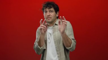Kıvırcık saçlı İspanyol bir adam canlı kırmızı bir arka plana karşı korkusunu dile getiriyor, ekose bir gömlek giyiyor ve elleri ve kolları çaprazlanmış bir savunma hareketi yapıyor..
