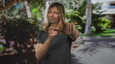 Uzun sarı saçlı bir adam ormanda elinde yeşil yapraklarla ezik işareti yapıyor, alay ediyor..