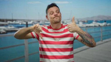Sahil limanındaki çizgili gömlekli genç adam, açık havada karışık duygular olduğunu düşündüren tekne ve su arkaplanlarına karşı dövmeleri olan baş parmaklarını hem yukarı hem aşağı gösteriyor.