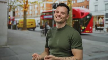 İspanyol adam kırmızı otobüsler ve binalarla çevrili bir şehir sokağında normal bir jest yapıyor, rahat giyiniyor ve kendine güvenini ve pozitifliğini ifade ediyor.