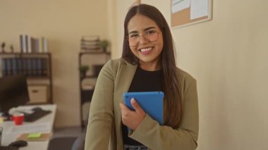 Ofis ortamında gülümseyen bir kadın, iş malzemeleri rafları ve masaüstü bilgisayarlarıyla çevrili tableti sıcacık bir şekilde tutarak profesyonel bir ortam sunuyor..
