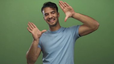 İspanyol asıllı genç adam, izole edilmiş yeşil arka plana gülümsüyor. Elleriyle çerçeveli jestler yapıyor. Neşeli bir ifade ve olumlu bir tavır sergiliyor..