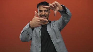 Genç İspanyol asıllı bir adam, elleri ile izole edilmiş kırmızı bir arkaplanda hareketler yapıyor, yaratıcılık ve odak noktasını bir film yapımı veya fotoğrafçılık bağlamında ifade ediyor..