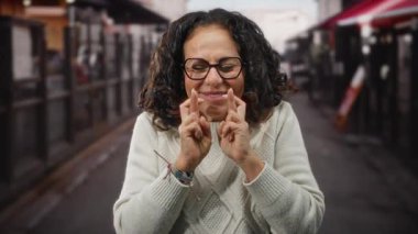 Kıvırcık saçlı, gülümseyen olgun bir kadın. Parlak bir günde, sıcak bir restoranın terasında krem rengi bir kazak ve gözlük giyerek dikilirken..