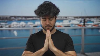 Young man with beard praying at port with ships in background, expressing serenity by the sea under clear sky.