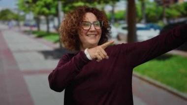 Woman pointing both directions in an outdoor park setting, showcasing a cheerful expression with brunette hair and glasses, surrounded by green trees and a cityscape.