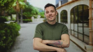 Yeşil gömlekli gülümseyen adam ağaçlarla ve binalarla dolu güneşli bir caddede kollarını kavuşturarak rahat ve kendine güvenen bir atmosfer yaratıyor.