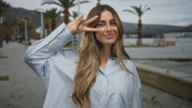 Sarışın genç bir kadın, palmiye ağaçlarıyla dolu bir marina güvertesinde, parmaklarıyla bir barış işareti tutuyor..