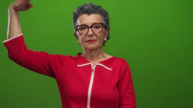 Gri saçlı yaşlı kadın canlı yeşil bir arkaplan önünde kırmızı elbise ve gözlük takarak selam veriyor..