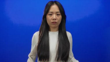 Belirsizliğini ifade eden, elleri mavi arka plan duvarında beyaz gömlek ve uzun saç giyen, kafa karıştıran ya da duyguları sorgulayan bir kadın..