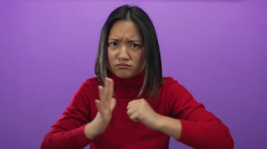 Güçlü duyguları ve odaklanmış ifadeyi vurgulayan kırmızı bir kazak giyen canlı mor arka plan karşısında kararlı olduğunu gösteren genç bir kadın.