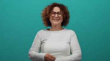 Orta yaşlı bir kadın, hayat dolu yeşil bir arka plana gülümsüyor, rahat beyaz bir kazakla güven ve mutluluk saçıyor, neşeli bir ifade sergiliyor..